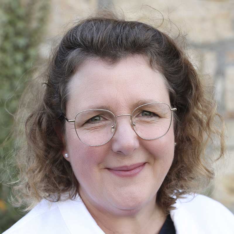 Eine Frau mit Brille und weißem Kittel lächelt in die Kamera. Sie hat mittellanges, lockiges Haar.