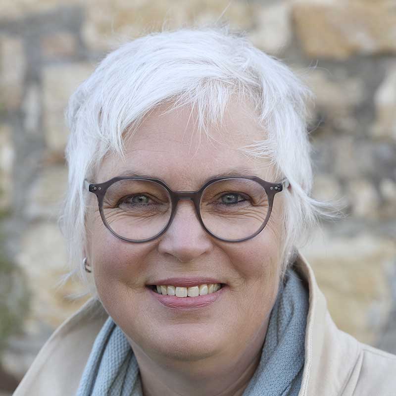 Eine Frau mit kurzen weißen Haaren und Brille lächelt.