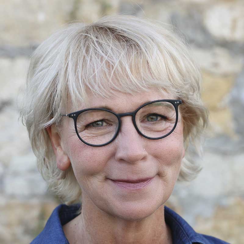 Eine Dame mit kurzen, blonden Haaren und schwarzer Brille lächelt.