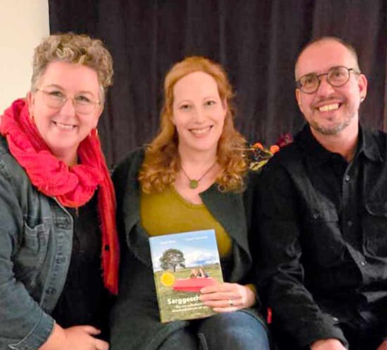 Ein Foto von drei Personen, die ein Buch halten. Die Personen lächeln.